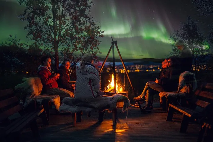 Guests sitting around a campfire under green northern lights at a husky camp