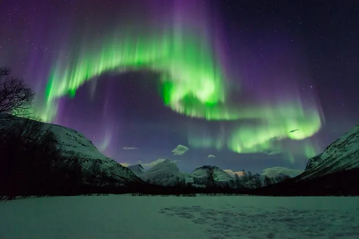 Grüne Nordlichter, die sich über einem verschneiten Tal mit dunklen Bergen wölben
